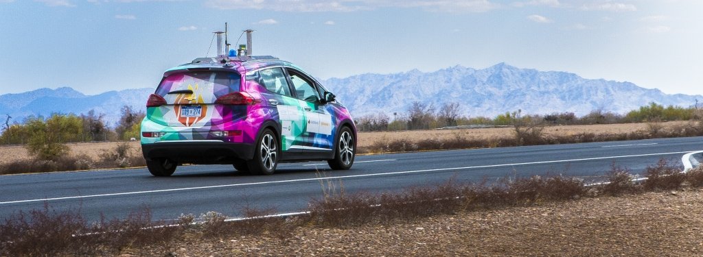 A car with LIDAR and GPS equipment on top drives on a highway with desert and mountains in the background. 