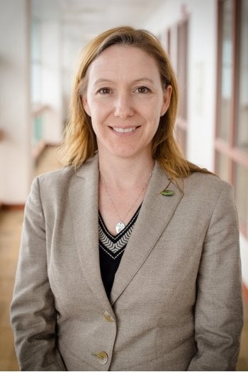 A woman in a suit coat stands in a brightly lit hallway.