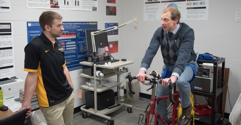 two men talking in a lab with monitors, fans, there are posters on the walls and one of the men is riding an exercise bike