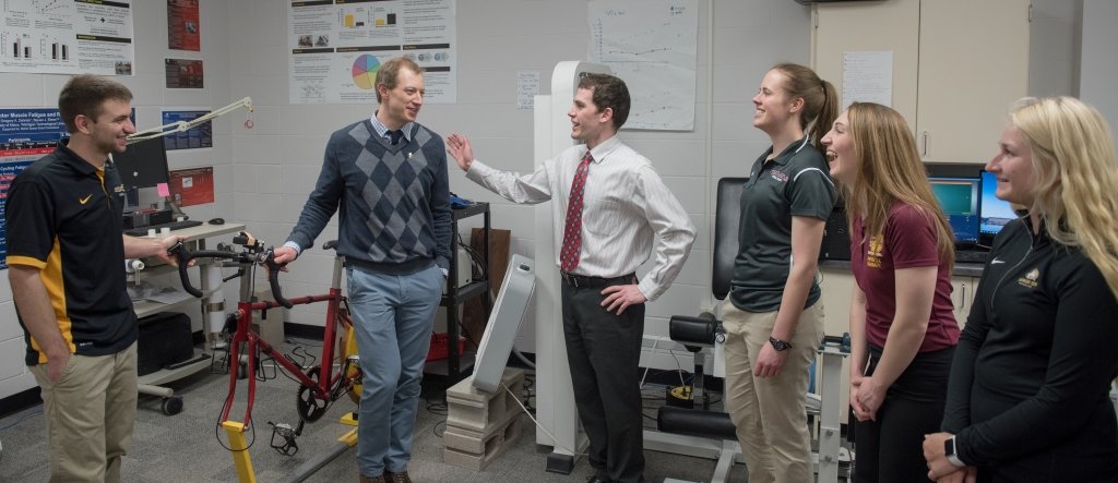 three men and three women talking in an exercise lab with a stationary bicycle, computer monitors, and posters on the wall 