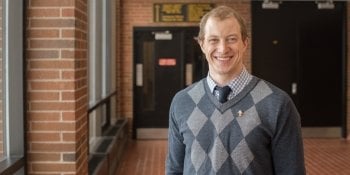 smiling man next to a brick wall with doors behind him wearing a diamond-patterned sweater and a tie