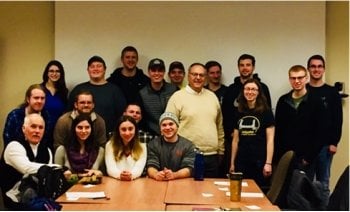 a group of male and female college students with front row seated and back row standing and their two male advisors in a conference room with a conference table in front of them.