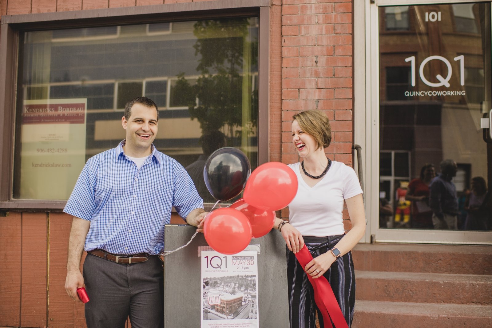 A man and a woman smile while standing next to a sign that reads 1Q1 launch party. There are balloons and each hold a ribbon.