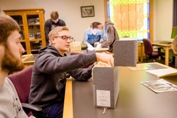 young male students look in gray file boxes with stained glass windows and other students doing research in a library archive space with file cabinets and white walls 