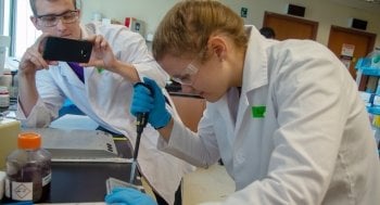 Two students in a lab. A male student takes a picture of the female student, who is pipetting, with his phone.