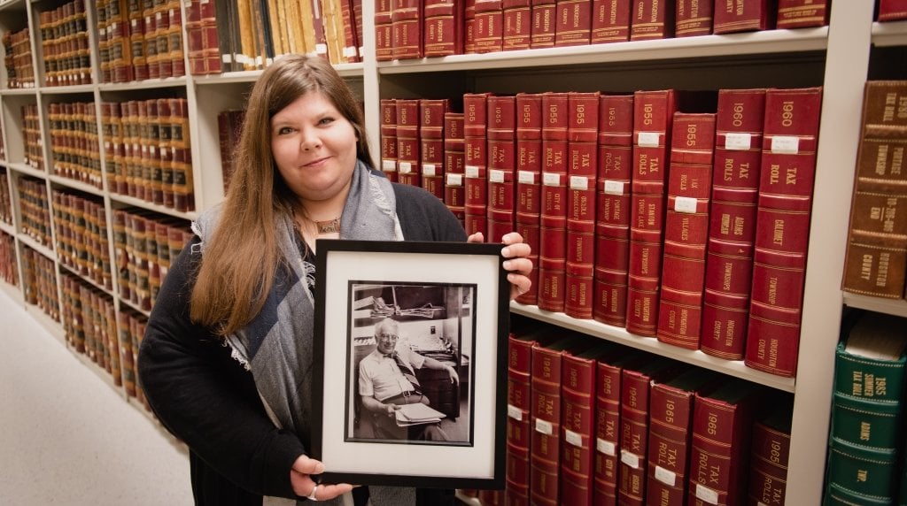 A woman with long hair holds a black and white framed photo of an older man with stacks of bound library volumes in the background