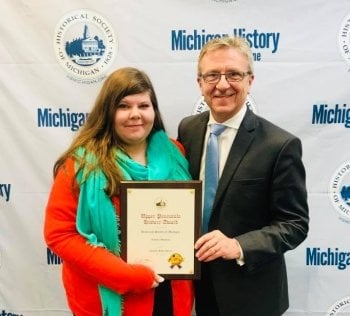 a woman and a man stand behind a Historical Society of Michigan Banner; she is holding a framed award