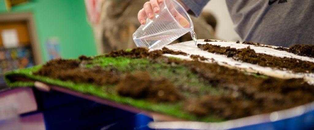 cup of water in a person's hand being poured over a pan of soil and green material with a green background in a science experiment in a classroom 