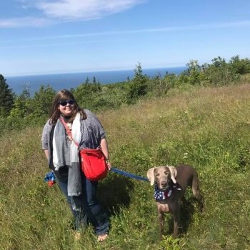 A woman and a Weimarainer dog on top of a grassy hill overlooking Lake Superior with blue water and blue sky