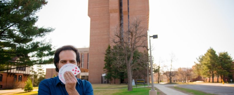 young man with cards fanned out in his hand standing on the grass next to a sidewalk on a college campus with a tall brick building behind him