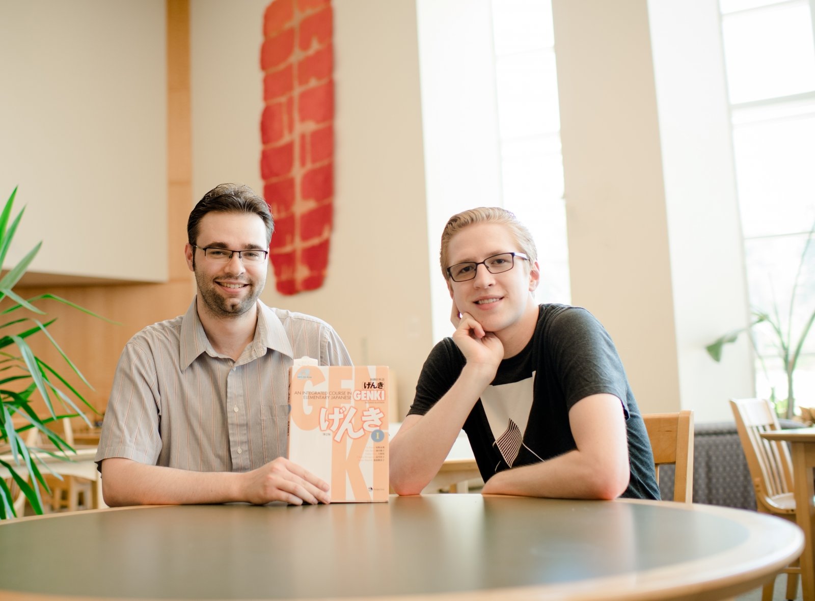 Two white male students sit at a round table. One holds up a book with the title elementary Japanese.