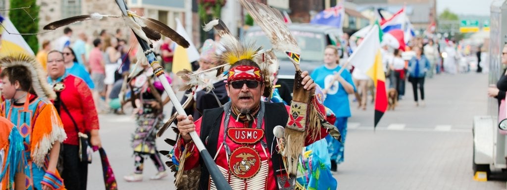 A man in native American regalia carrying eagle feathers marches in a parade with a young man and woman also in Native American regalia in the background, and lines of people walking in the street, carrying national flags and people on the side of the street outside watching the parade 