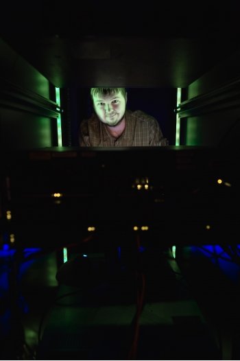 A young male student peers out between computer servers in a dark IT lab lit with green