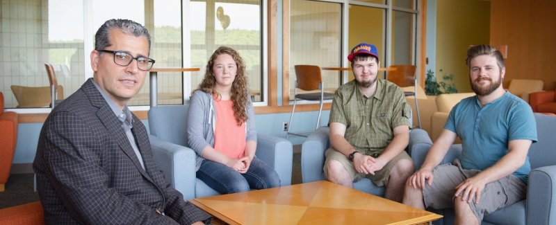 A professor, male, with a male, female and male student seated on chairs in a student study lounge with natural light coming in from the windows