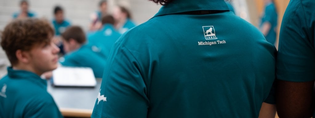 A Michigan Tech logo in white on the back of a teal shirt in the foreground with a side profile of one young man and eight other young people wearing teal polo shirts that match