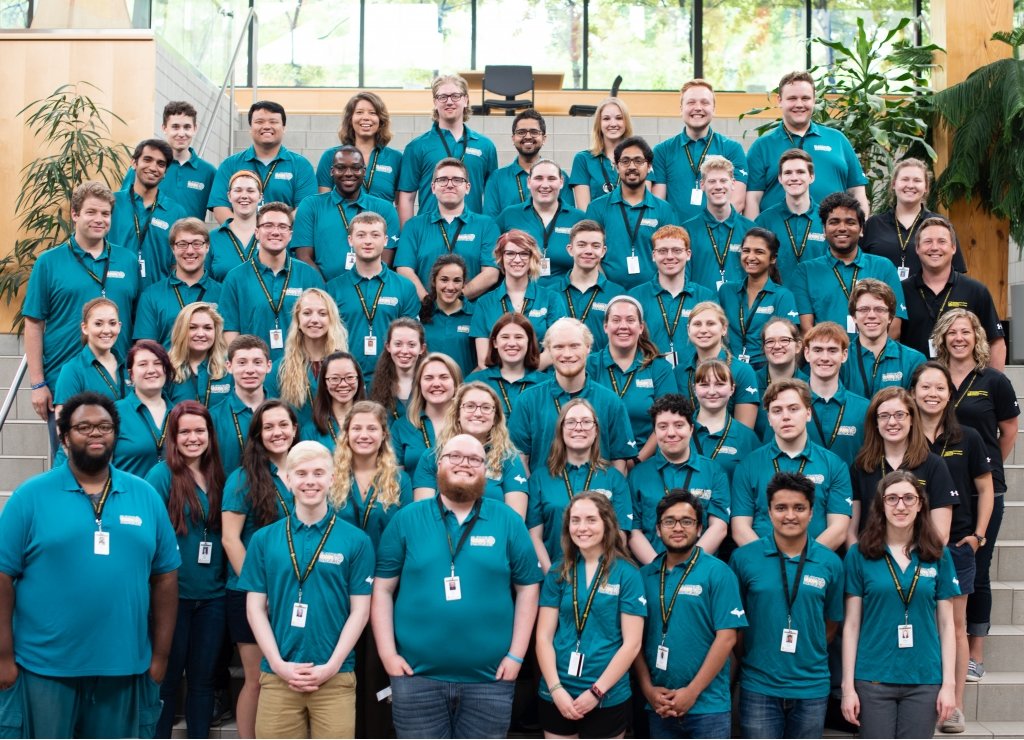 60 people in teal and black polo shirts standing on steps in rows smiling as a group