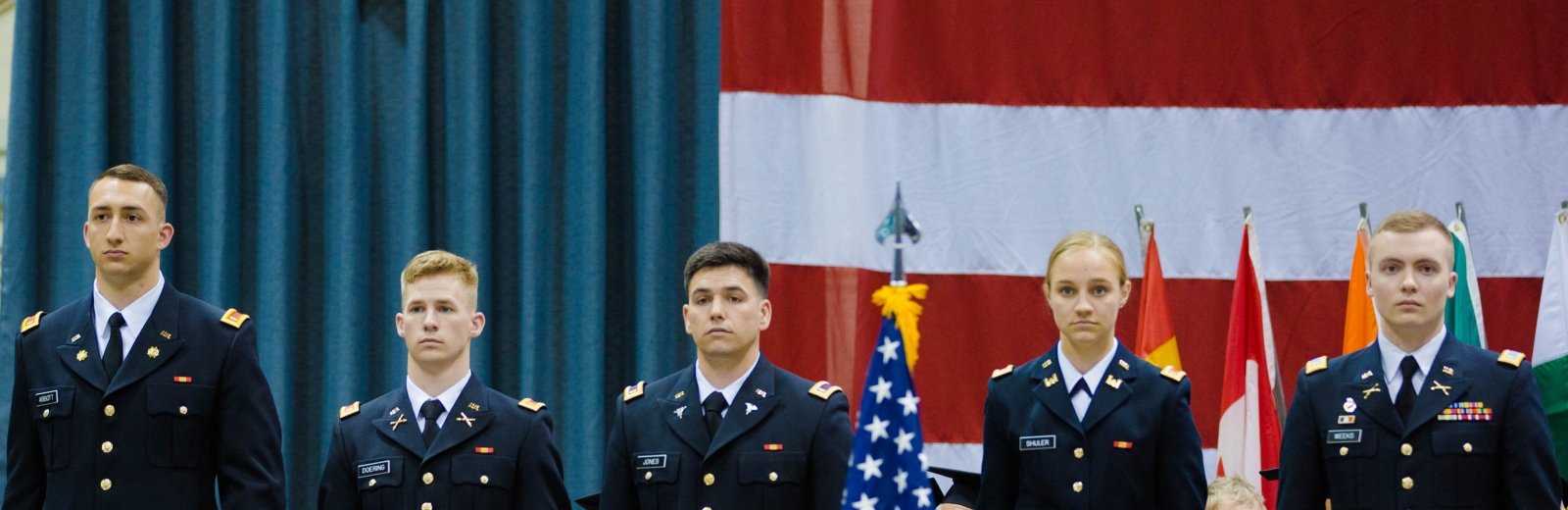 Four Army ROTC cadets and one Air Force ROTC cadet stand at attention on stage at commencement. American flag in background and foreground. Headed caps of graduates in audience in foreground too.