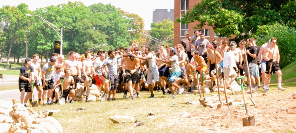 100 male football players in shorts and an older female professor gesture excitedly on a grassy hill in front of shovels, sand and a sandbag with college campus brick buildings and large green and leafy trees in the background