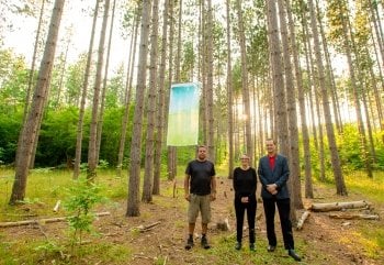 Two men and a woman stand beneath a cloth banner in a pine tree grove.