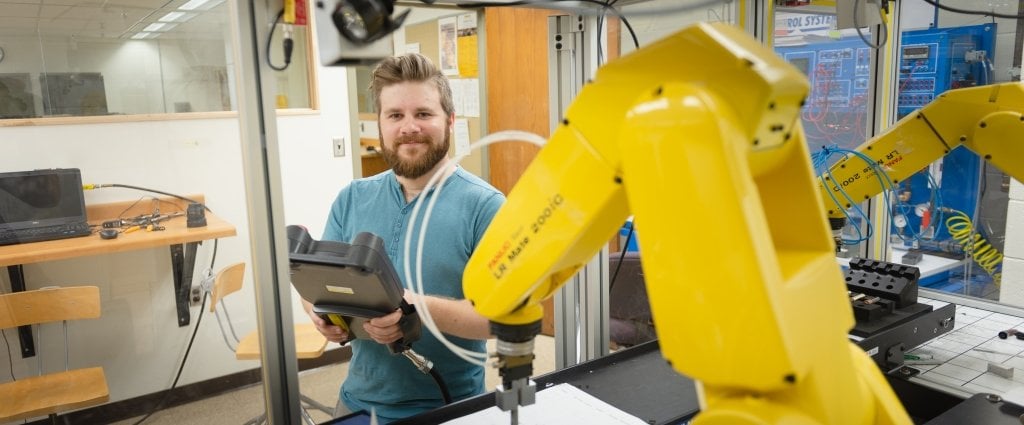 student holding a control panel stands next to a yellow robotic arm 