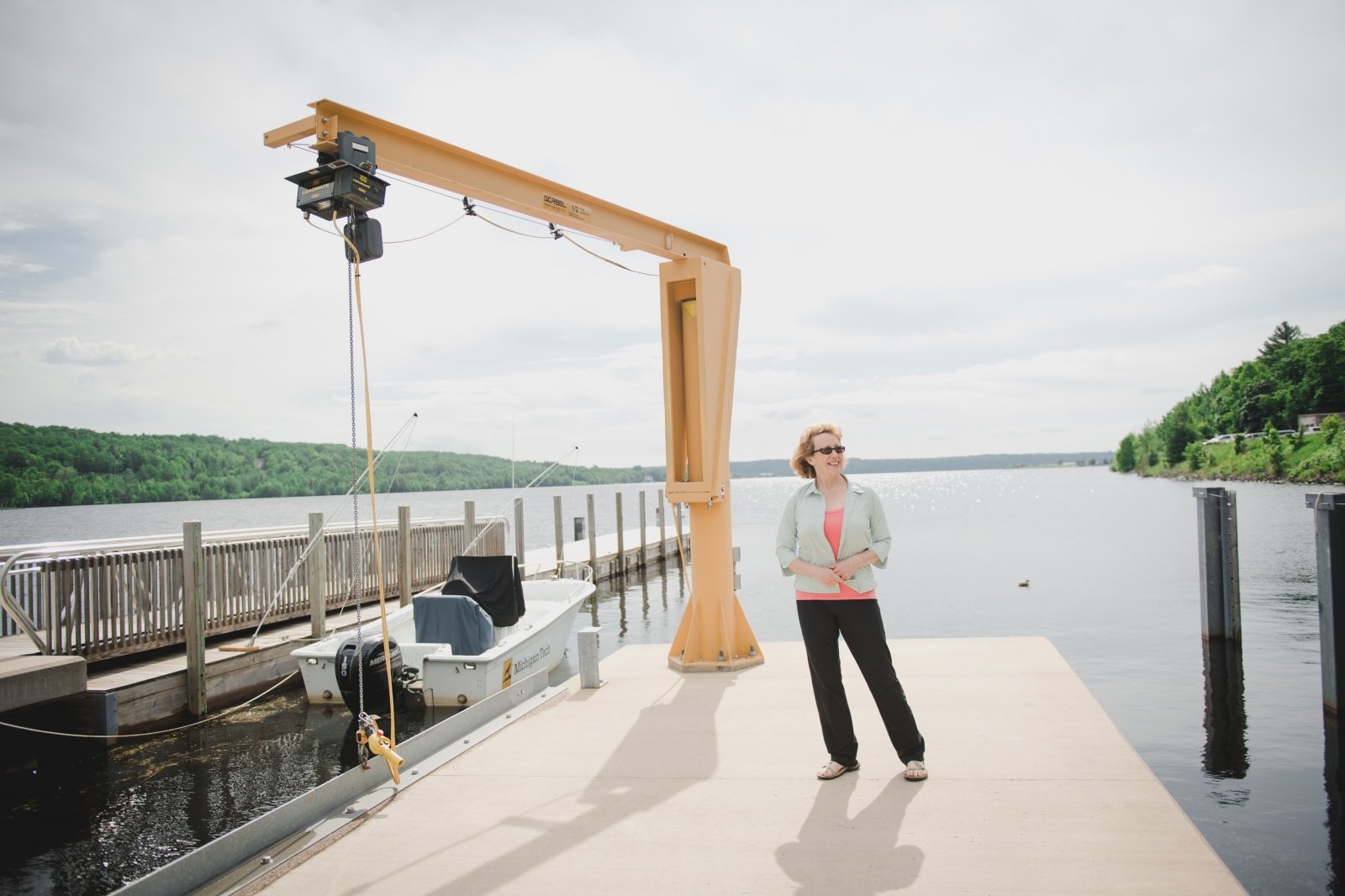 A woman standing on a dock next to a yellow boat lift with a canal in the background and forested hills. There is a boat with an outboard engine tied to the adjacent pier.     