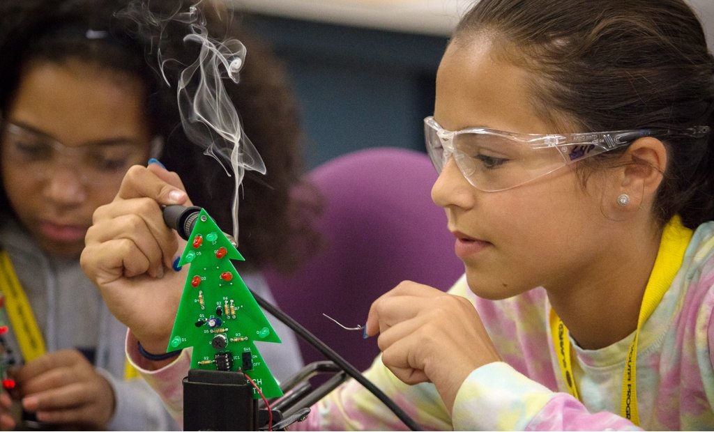 two young girls working on green pine-tree shaped circuit boards, wearing safety glasses, with the girl in the foreground using a soldering iron. 