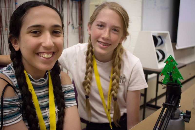 Two young girls with braids smiling at a summer camp in an interior location with a white wall.