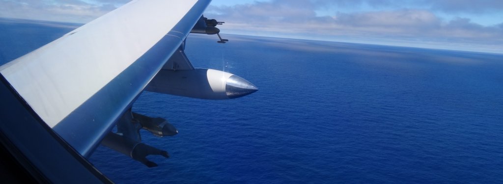 An airplane wing fitted with instruments, including the HOLODEC, flying over the ocean with clouds on the horizon.