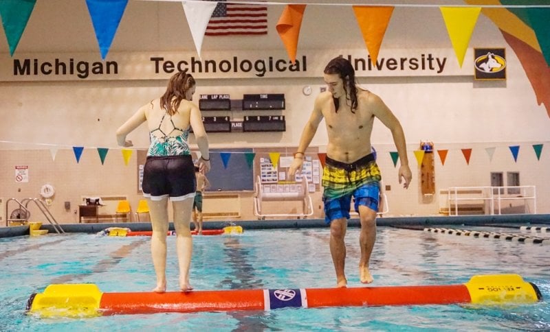 A female and a male student stand on a red log in an indoor swimming pool that's regulation size. Her back is to us, he is facing us, they are looking at each other's feet as they roll the log.