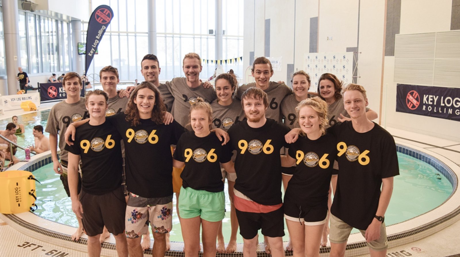 Students stand in a group wearing 906 T-shirts and shorts and bathing suits in front of an indoor pool at a log-rolling competition.