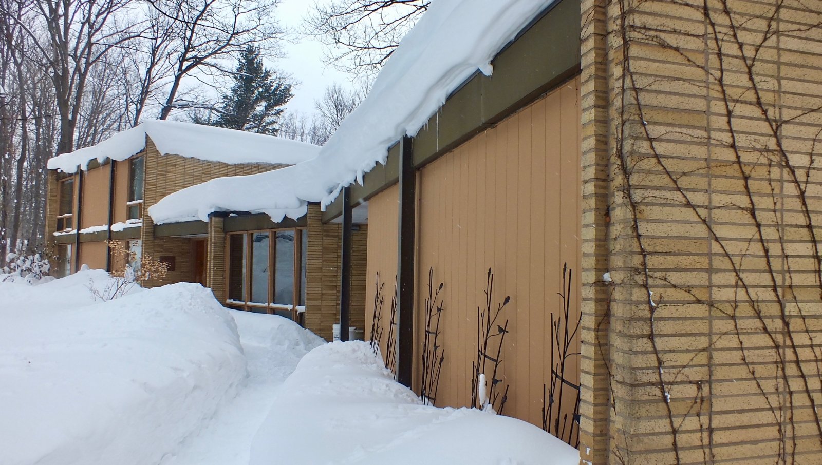 A tan brick and wood tri-level home with several feet of snow on the ground and bare trees in the background in winter. 