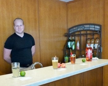 A young man stands behind a wooden bar with wood paneling in the background with soft drinks, glasses and fruit on the bar.