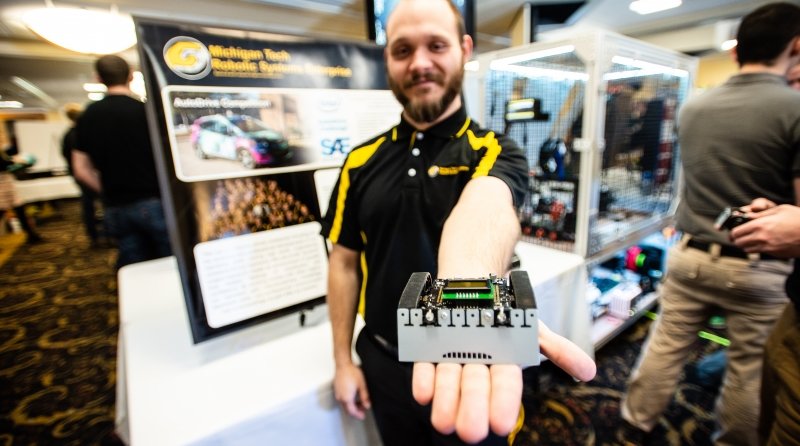 A young man in black and gold holds out a small circuit board at an Expo with display tables and people in the background
