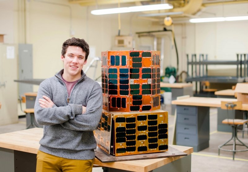 A young man in a lab classroom inside with crossed arms standing by a satellite with reflective panels sitting on a table, it's shaped somewhat like a large cake and is a little taller than a mini-fridge