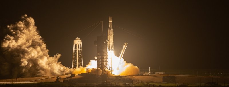 smoke and flames as a rocket launches from a NASA space pad with the SpaceX water tower in the background at night