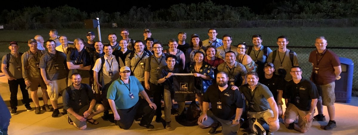 30 people in four rows with the front row kneeling stand on a concrete pad at night with a fence and kudzu in the background. Two of the people are holding a Michigan Tech flag.
