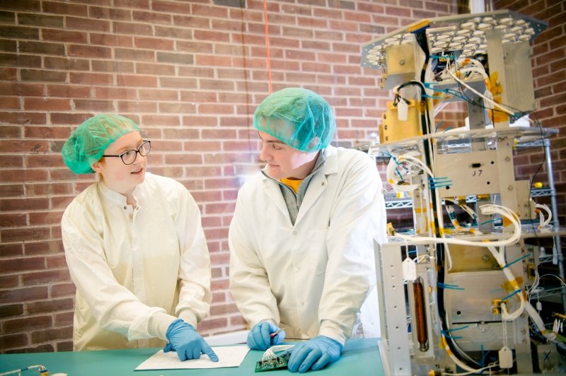 A young woman and a young man wearing scrubs, gloves, and caps in a clean room where a satellite is being assembled