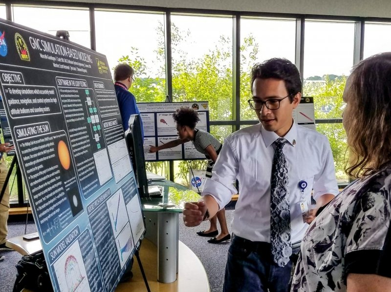A young man stands before a poster gesturing animatedly to a woman seen in profile at a presentation with the outdoors seen in the background through a glass wall, and a young woman giving a poster presentation in the background.