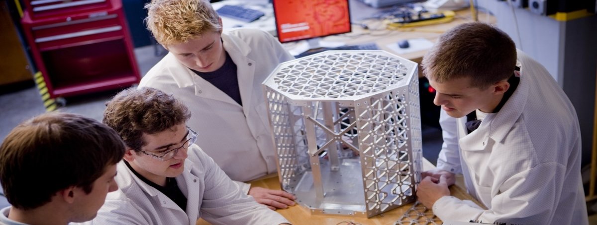 four students in white coats work on a circuit board and metal framework in an aerospace class in a lab wearing white coats