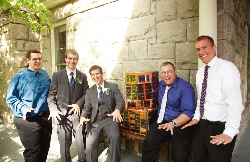 Five men in weddig attire, including two in tuxedos, show jazz hands in a sunny courtyard with a satellite on a table and a gray stone wall, white pillar and green tree in the far upper left, in the background. They are all smiling.