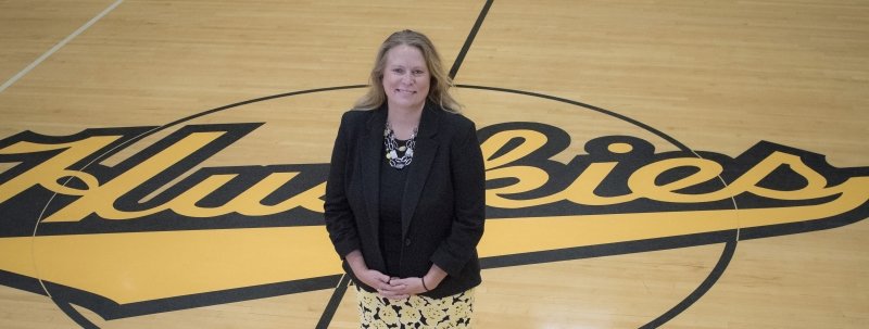 A woman in a suit smiles on a gym floor with a Huskies black and gold logo behind her.