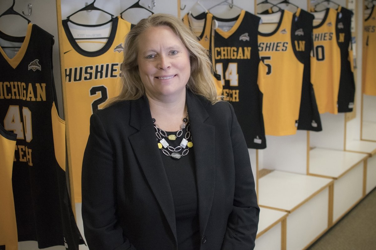 A women stands in a locker room with tank top uniforms behind her hanging from lockers that says Michigan Tech Huskies