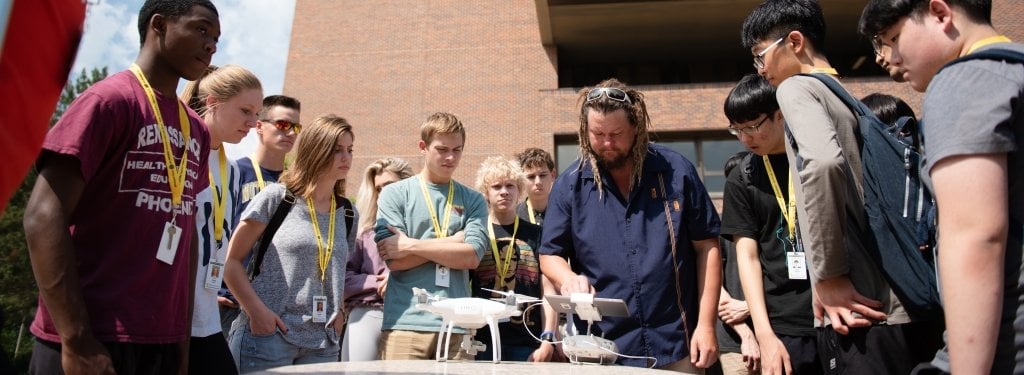 A group of students gathers around James Bialas, who has an aerial drone and drone controller in front of him.