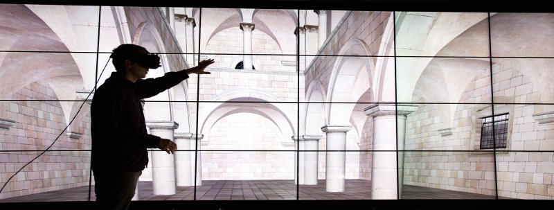 A person wearing a virtual reality headset motions his hands in front of a wall of computer screens