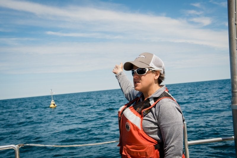 A scientist wearing white sunglasses stands on the deck of a ship gesturing to a black-and-gold buoy in the background on the water on Lake Superior.