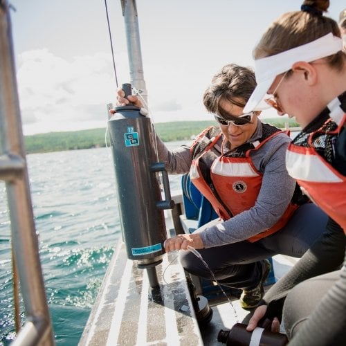 a woman researcher in an orange vest shows a student with a visor on how to empty out a Niskin bottle while conducting research on the deck of a ship.