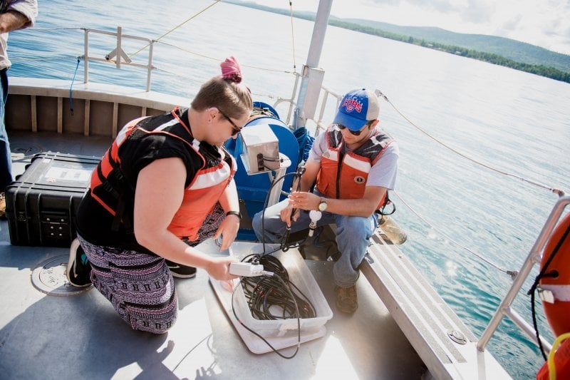 two students kneel on the deck of a boat wearing life jackets using light penetrating equipment.