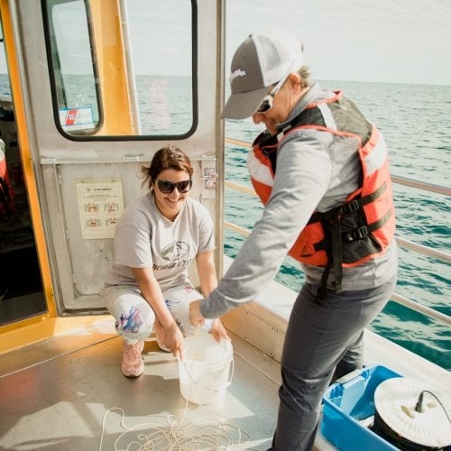 A woman stands on deck holding a line and another squats on the deck of a boat holding a line in a bucket on a ship in the blue waters of Lake Superior.