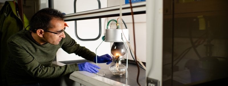 A man looks at a kerosene lamp hooked up to a vacuum under a laboratory vent hood.