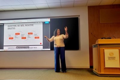 A woman on a stage gestures while giving a talk to Thomson Reuters news service with predictive analytics, algorithms and big data on the board behind her.